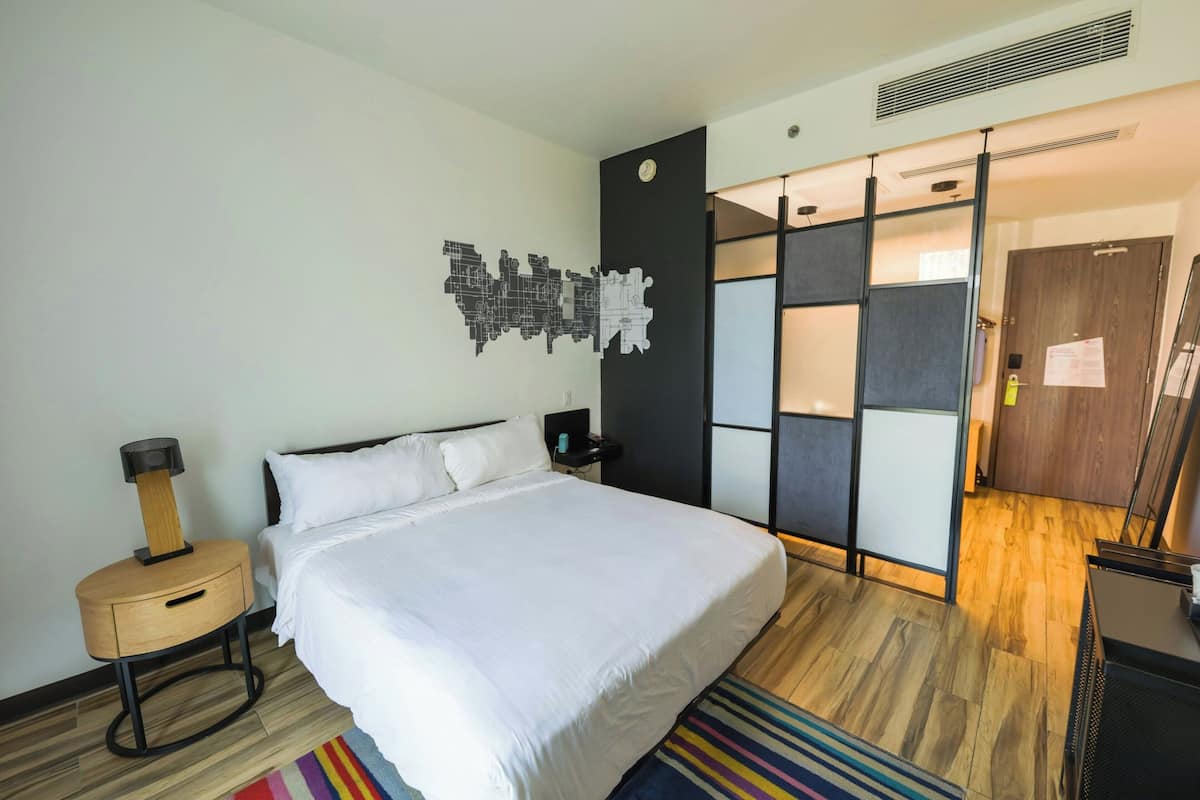 A modern hotel room with a large bed, a bedside table with a lamp, a wall clock, and a sliding door closet.