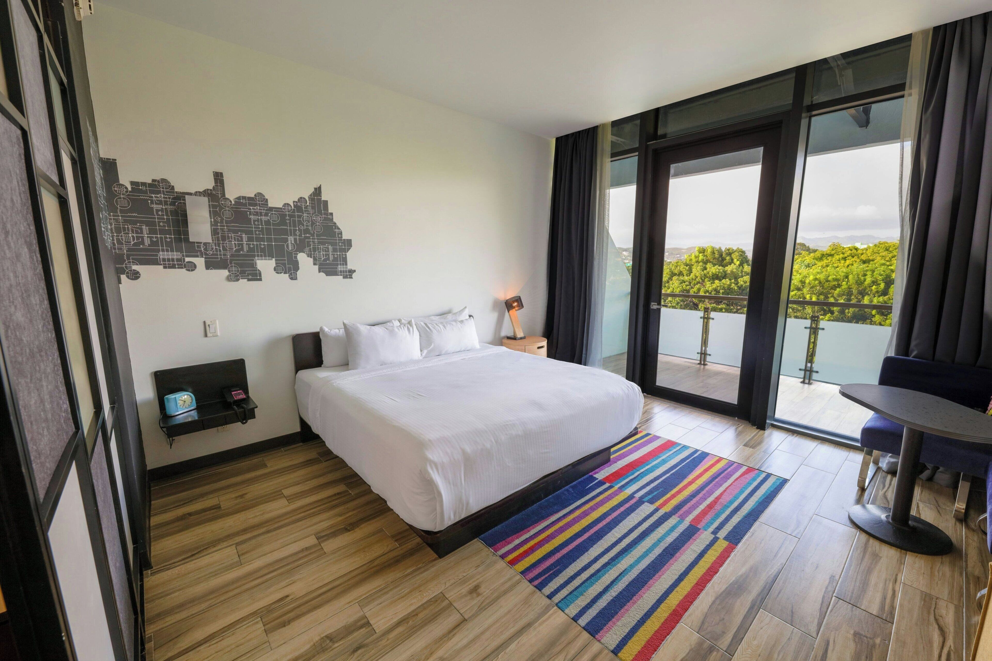 A modern hotel room with a large bed, a cityscape wall art, a balcony with a view, and a small table with a chair.