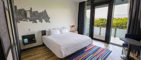 Una habitación de hotel moderna con una cama grande, un cuadro de arte urbano en la pared, un balcón con vistas y una pequeña mesa con una silla.