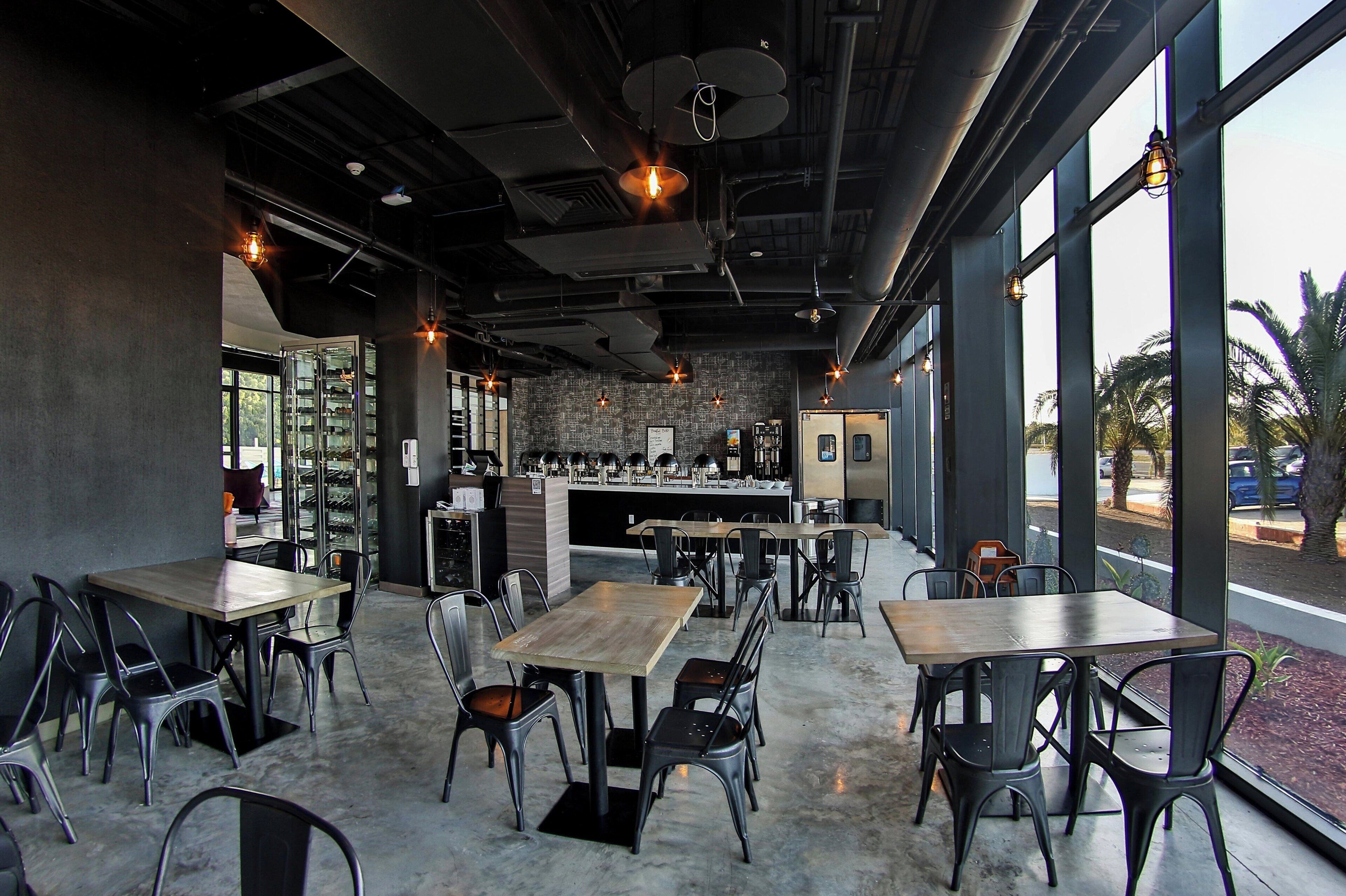 A modern cafe interior with tables and chairs, a bar area, and large windows offering a view of palm trees.