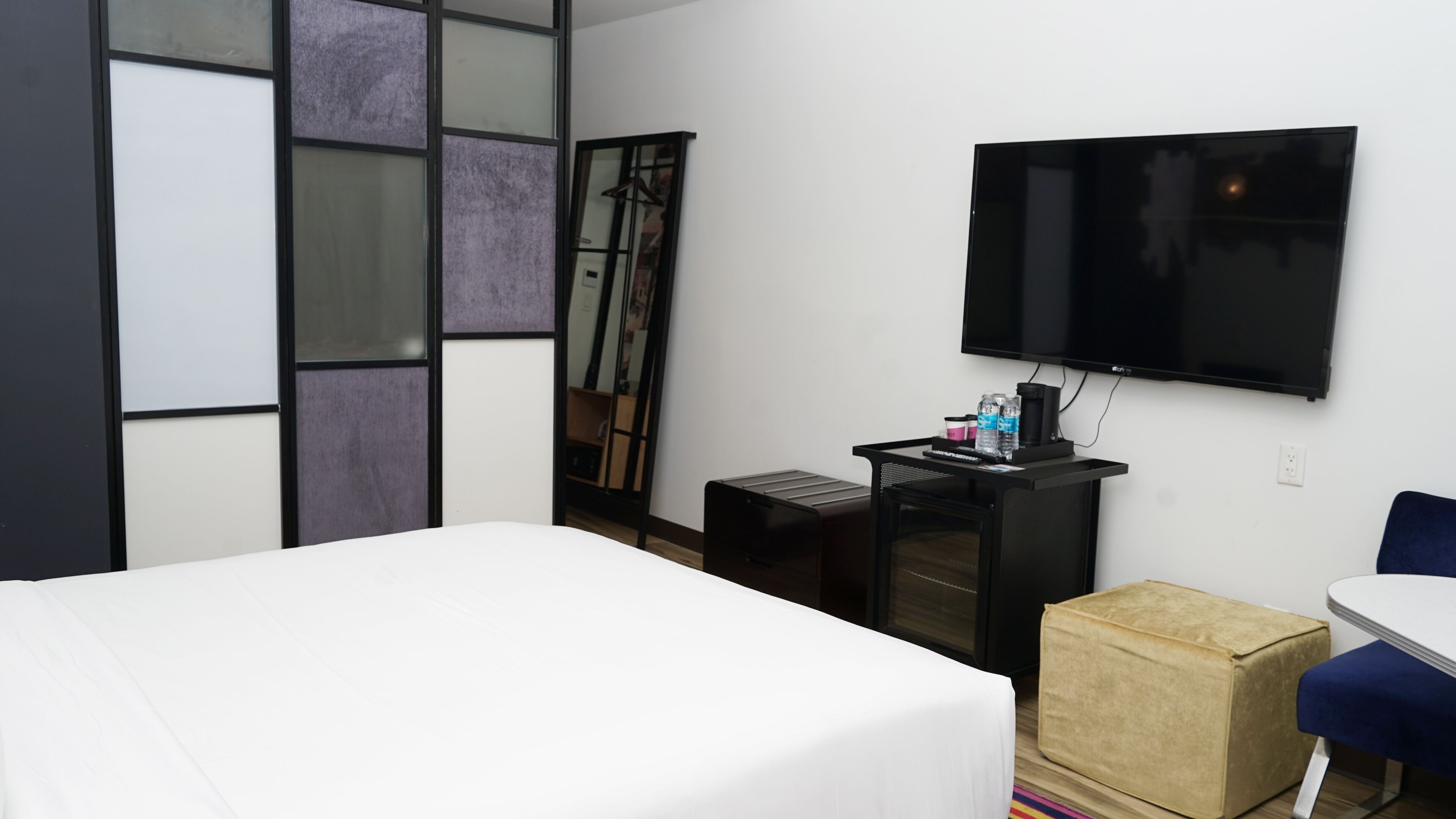 A modern hotel room with a large bed, a flat-screen TV mounted on the wall, a small desk with a computer and a coffee cup, and a blue chair.