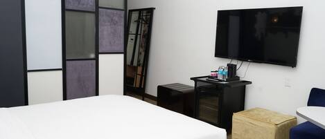 Habitación de hotel moderna con una cama grande, un televisor de pantalla plana en la pared, un escritorio pequeño con una computadora y una taza de café, y una silla azul.
