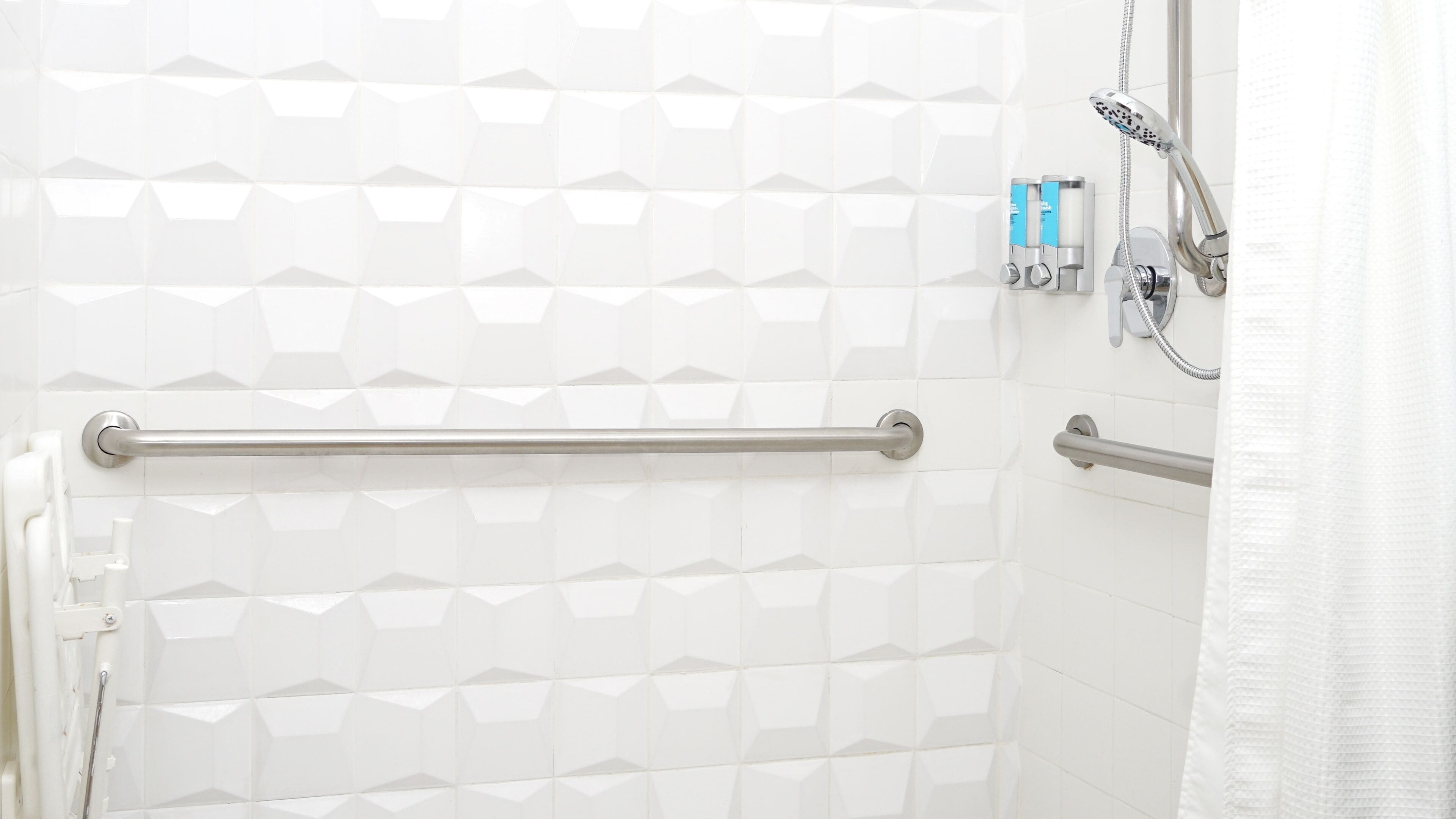 Zona de ducha con paredes de azulejos blancos, barra de apoyo, cabezal de ducha manual y un perchero para toallas.