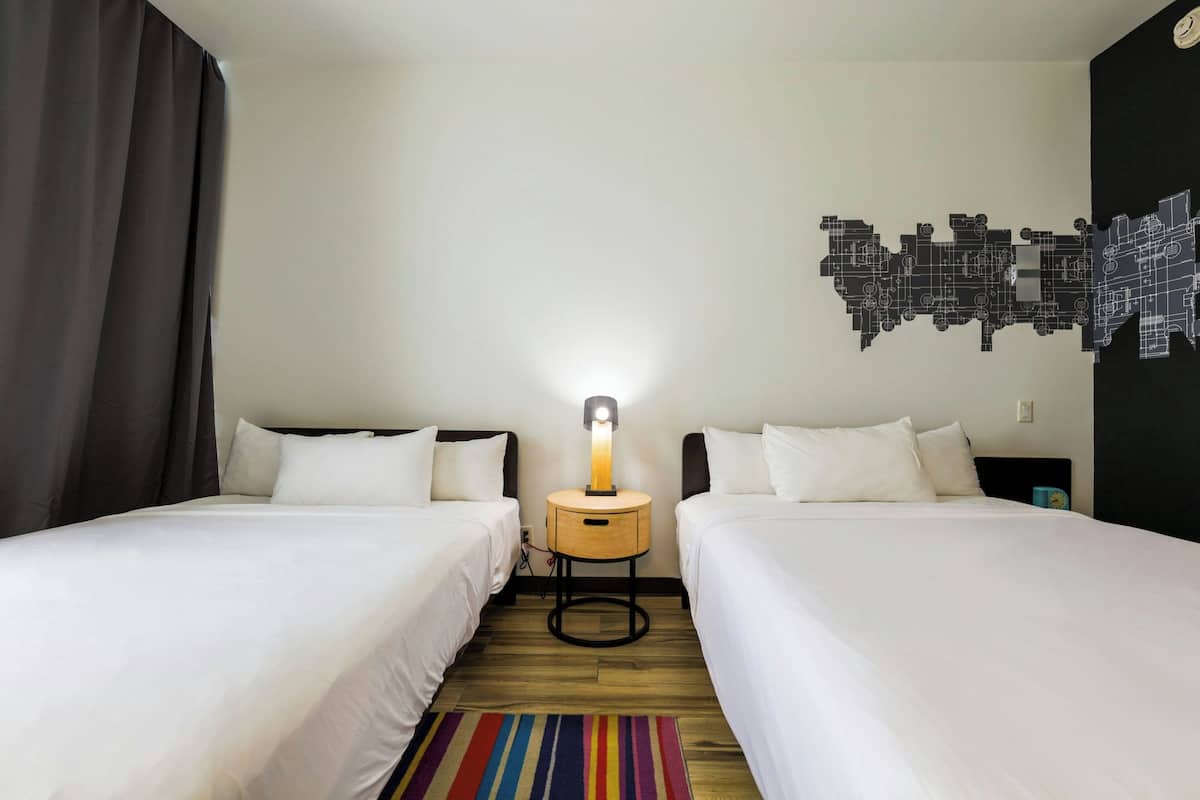 A hotel room with two single beds, a nightstand with a lamp, a colorful striped rug, and a wall art piece.