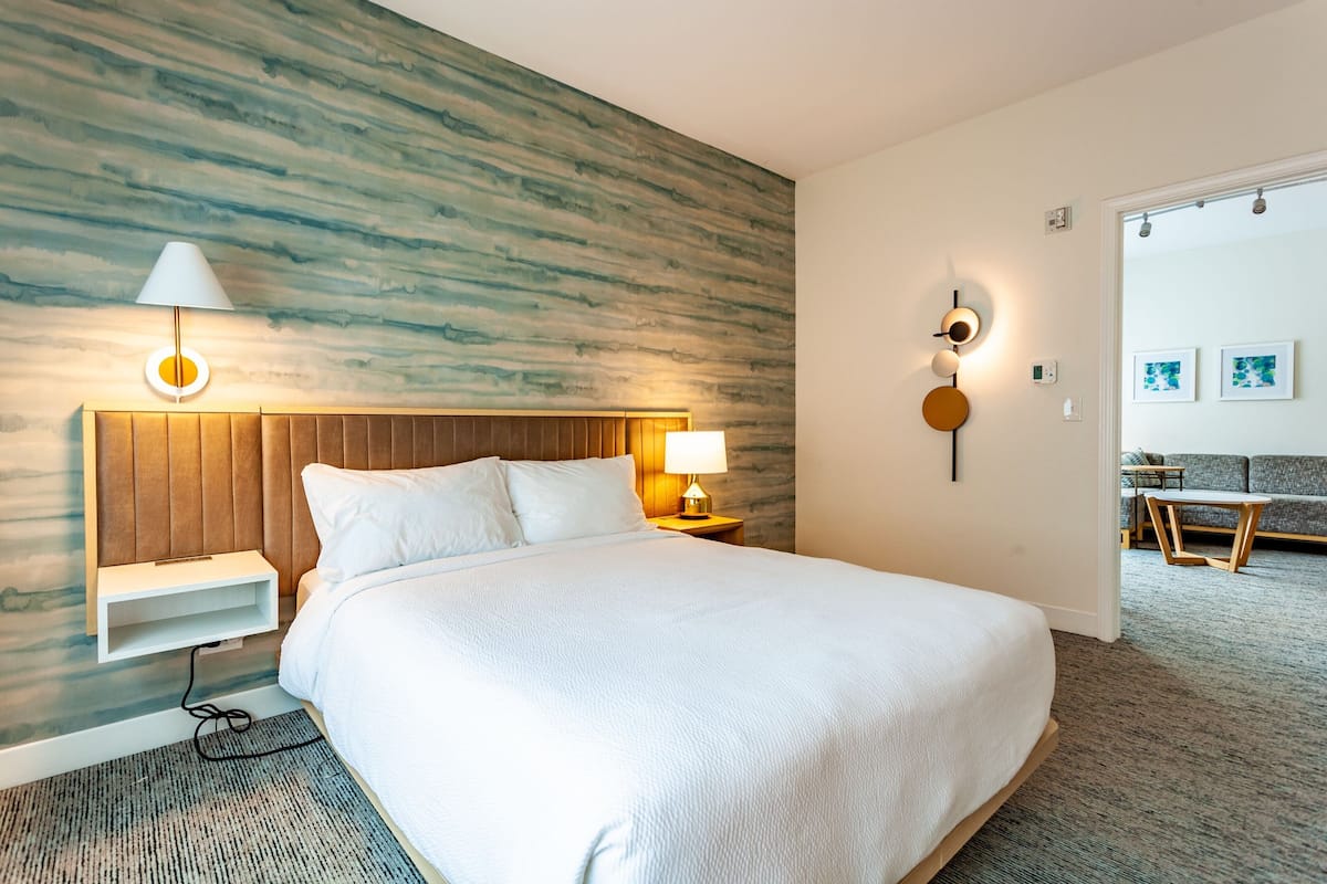Ein modernes Hotelzimmer mit einem großen Bett, einem Holz-Kopfteil, Nachttischen mit Lampen und Blick in den Wohnbereich.