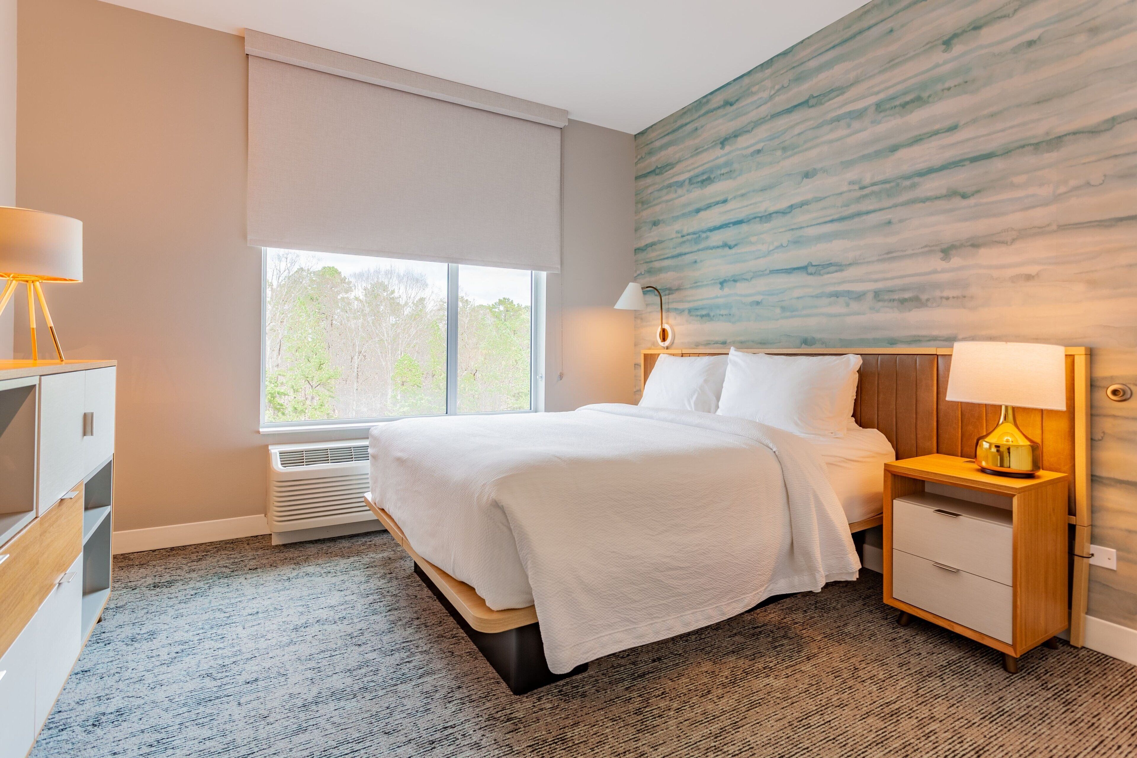 A hotel room with a bed, bedside tables, a lamp, a heater, and a large window.