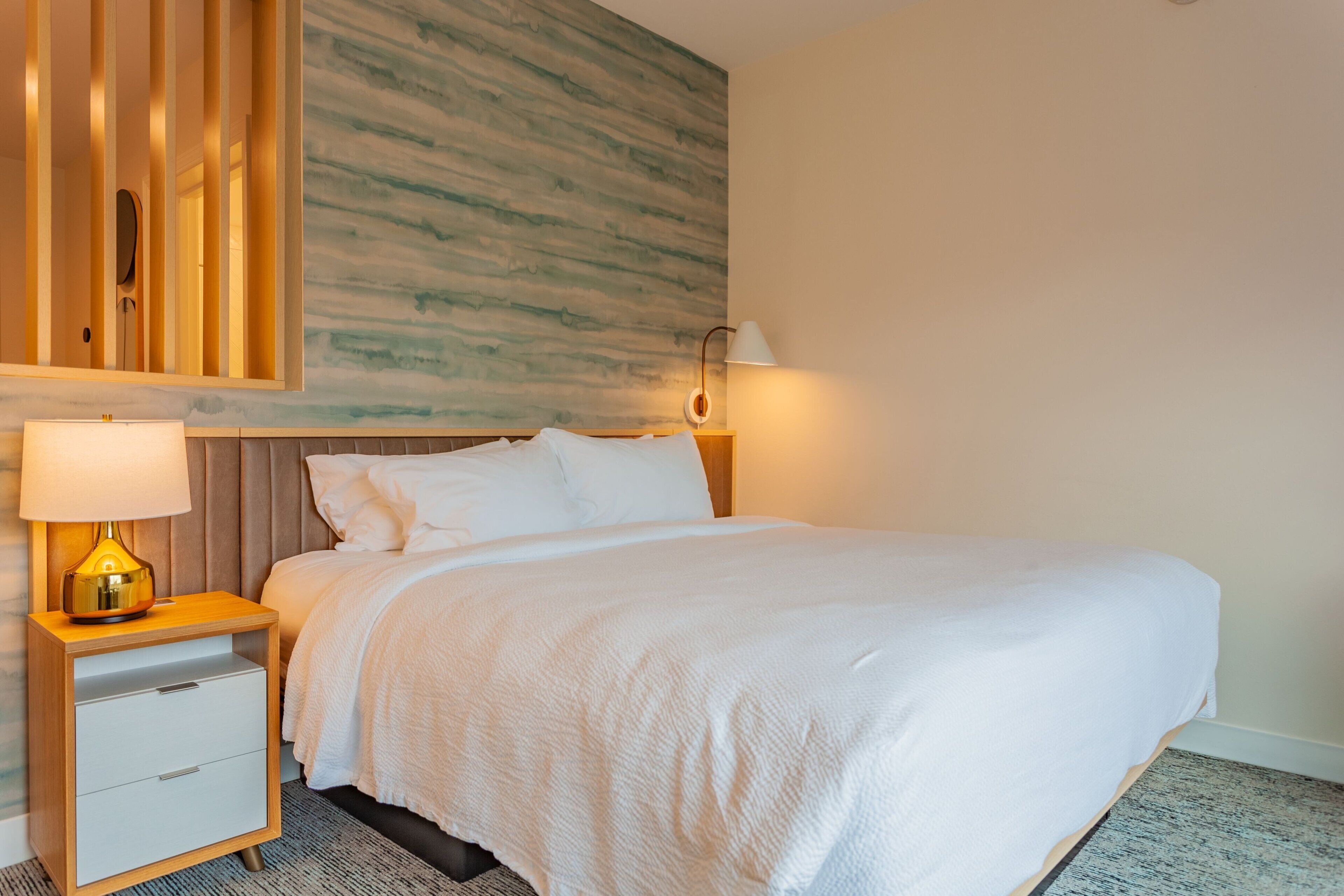 A neatly made bed with white linens, a wooden nightstand with a lamp, and a wall with a textured pattern.