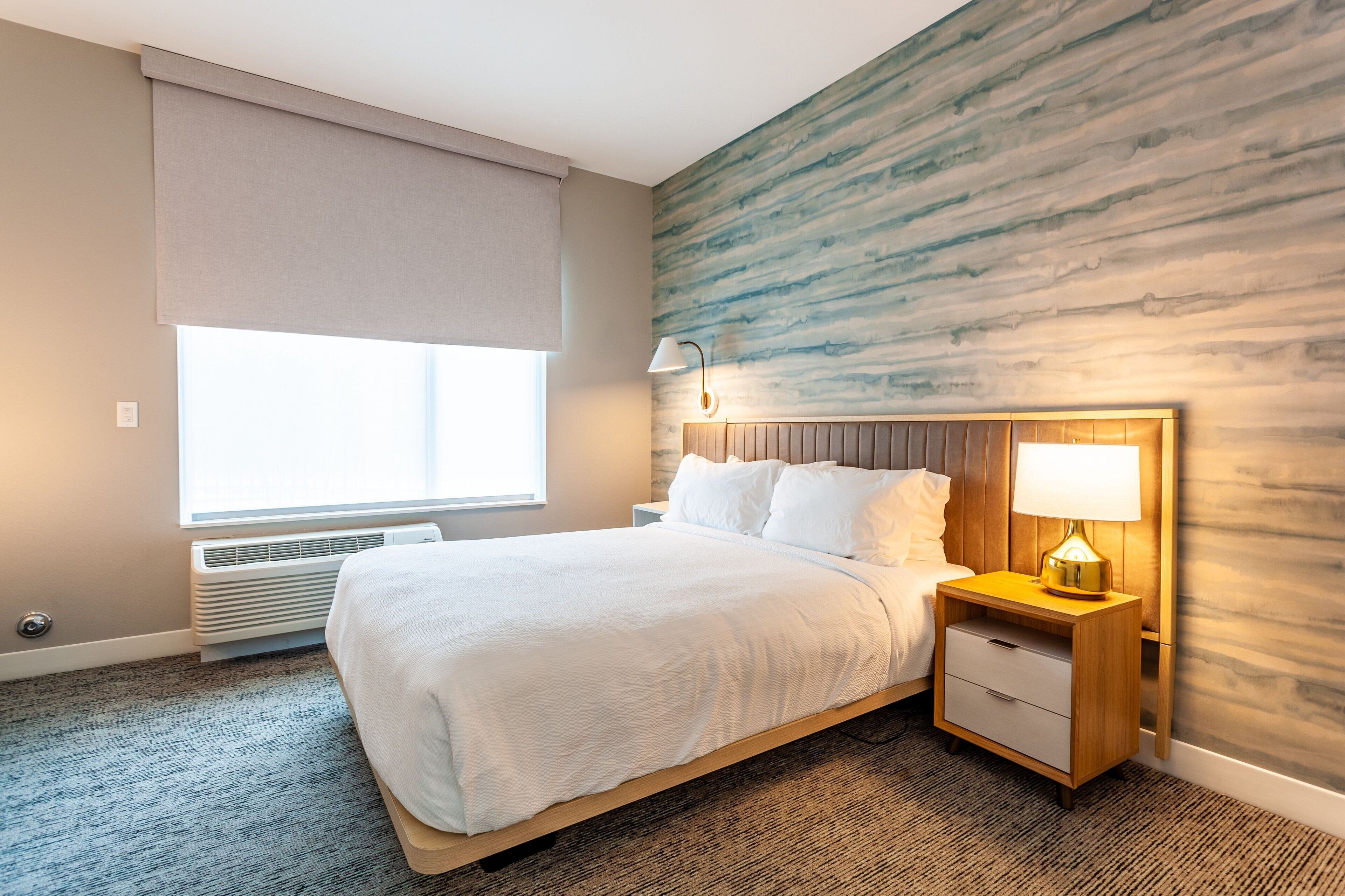 A hotel room with a bed, a nightstand, a lamp, a window with a blind, and a wall with a wood-like texture.