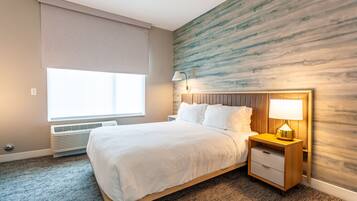 A hotel room with a bed, a nightstand, a lamp, a window with a blind, and a wall with a wood-like texture.