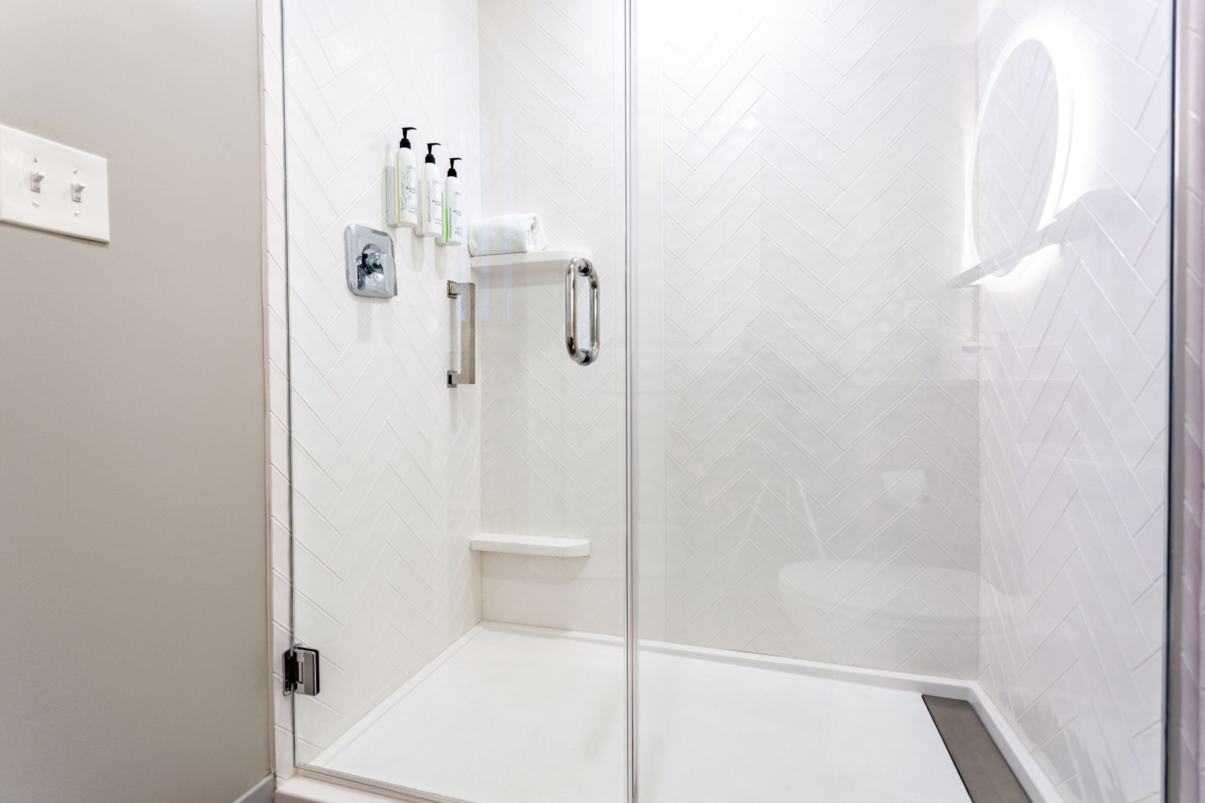 A modern shower area with a glass door, white herringbone tiles, a handheld showerhead, and a shelf with toiletries.