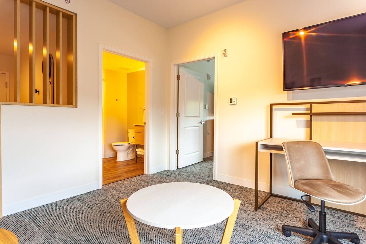 Ein modernes Zimmer mit einem runden weißen Tisch, einem Drehsessel, einem flachbildfernseher an der Wand und einem Raumteiler aus Holzlatten.