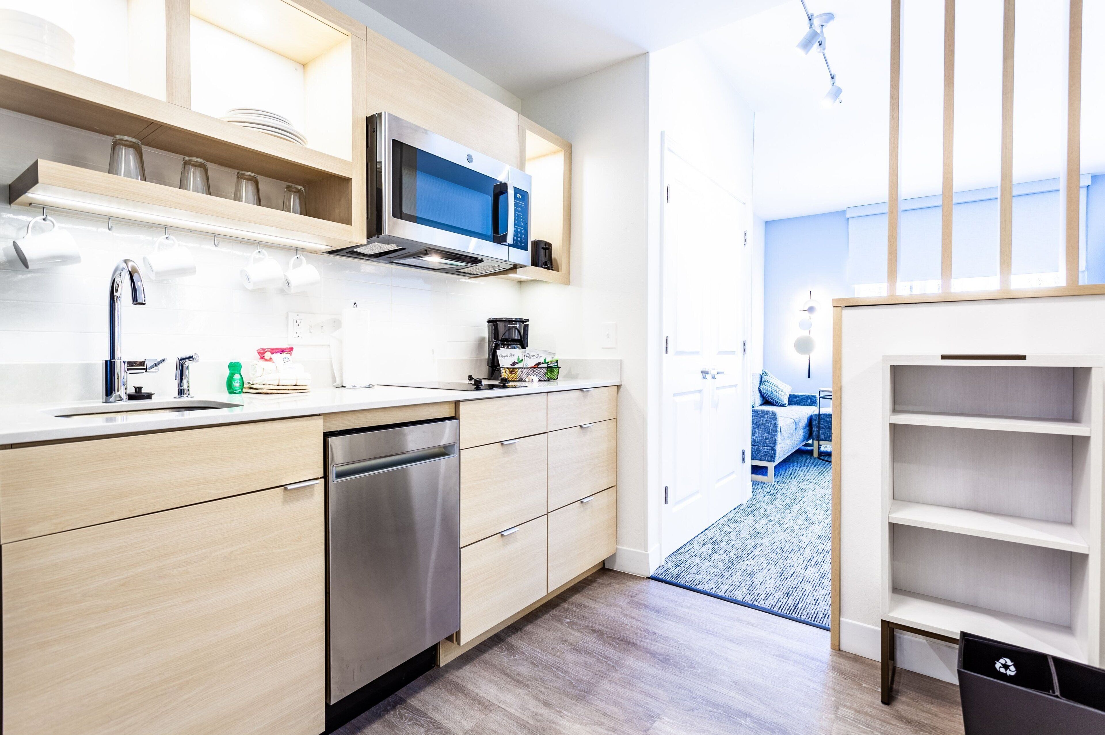 A modern kitchen with a dishwasher, microwave, and coffee maker. There is a wooden shelf above the counter and a view into the living area through an open door.