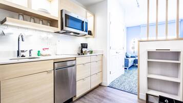 A modern kitchen with a dishwasher, microwave, and coffee maker. There is a wooden shelf above the counter and a view into the living area through an open door.