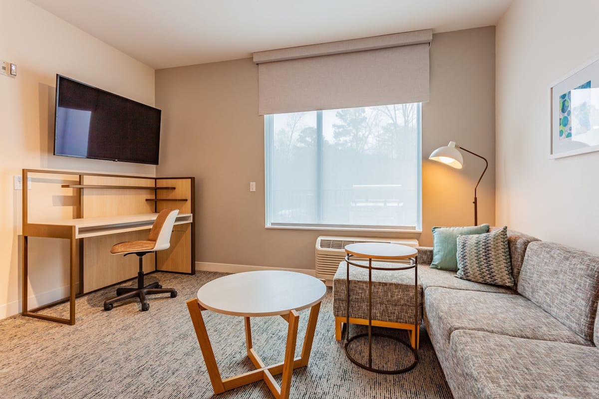 Ein modernes Hotelzimmer mit einem an der Wand befestigten Flachbildfernseher, einem Schreibtisch mit Stuhl, einem runden Tisch, einer Couch und einer Stehlampe.