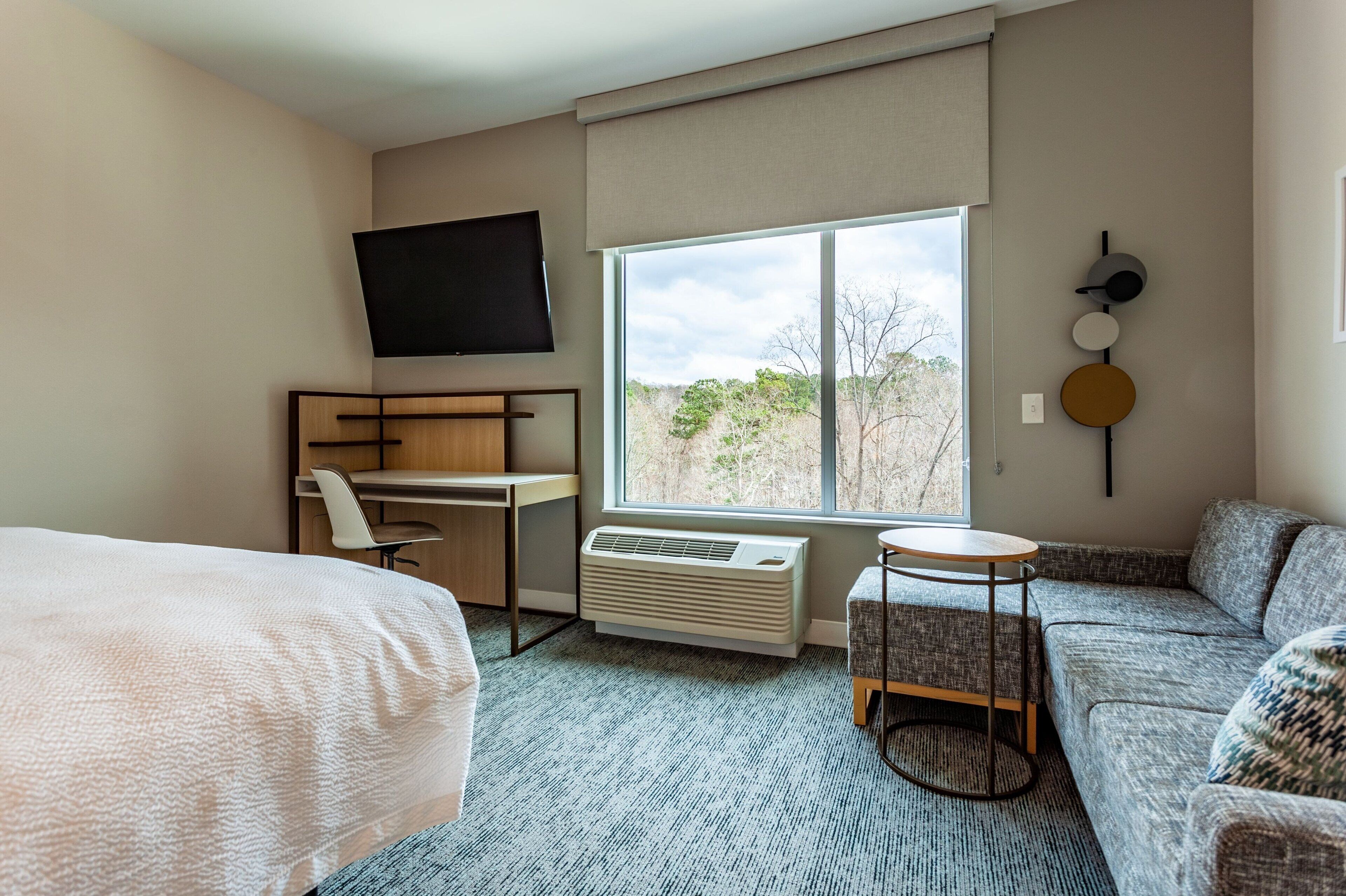 A hotel room with a bed, a desk, a chair, a sofa, a small table, and a large window with a view of trees.