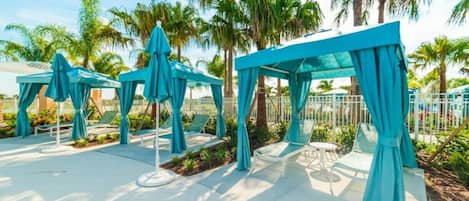 Relax in a serene outdoor space with lounge chairs and tables under blue canopies, surrounded by a white fence and lush greenery.