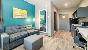 A modern hotel lobby with a teal accent wall, a grey sofa, a small ottoman, a kitchen area with a sink and countertop, and a mirror reflecting the space. - Everglades Adventures Hotel Suites by Ivey House (Everglades City)