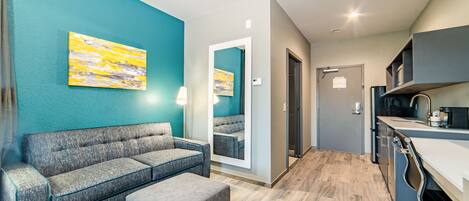 Un moderno vestíbulo de hotel con una pared de acento color verde azulado, un sofá gris, un pequeño reposapiés, una zona de cocina con un lavamanos y encimera, y un espejo que refleja el espacio.