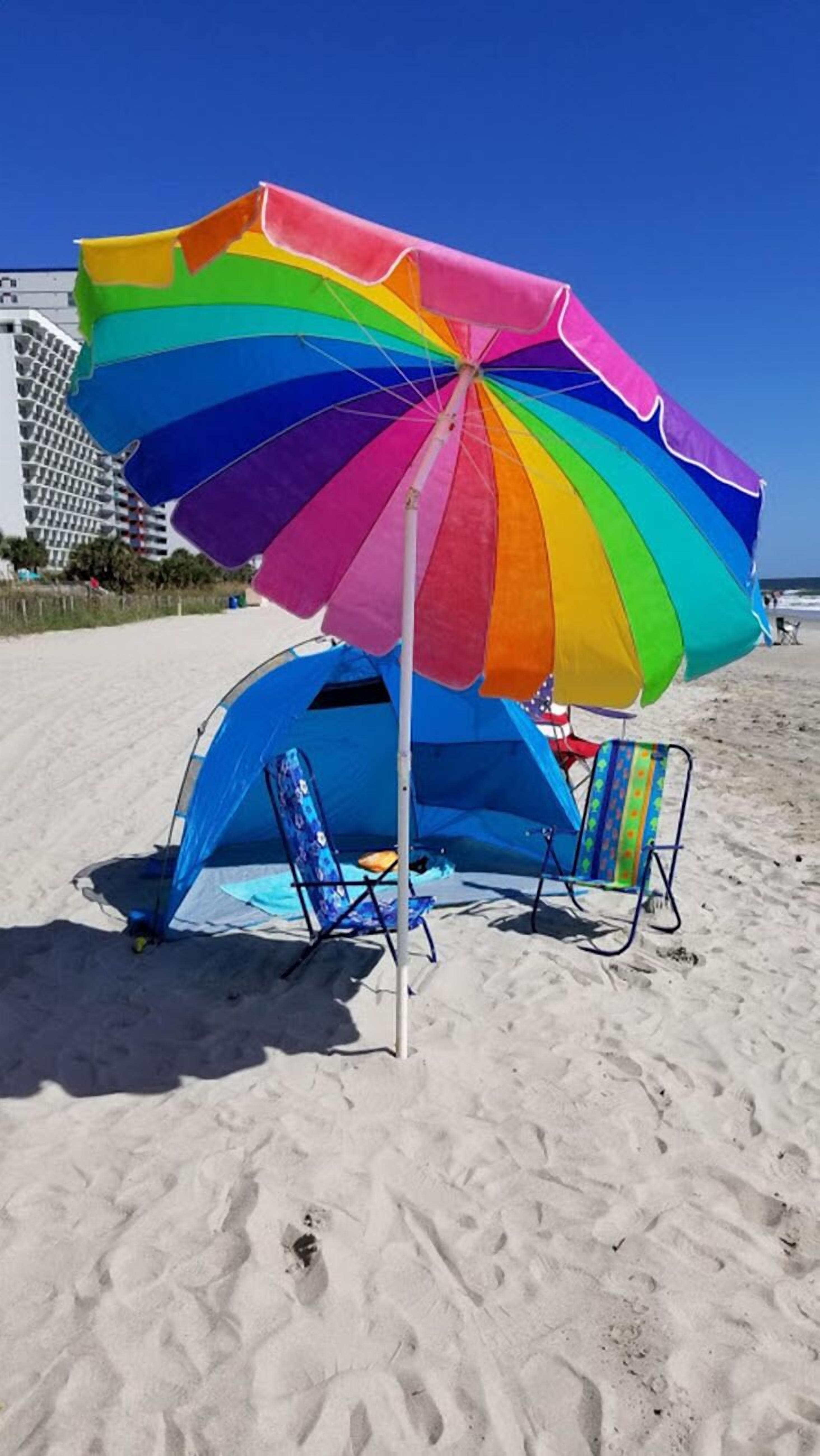 Una carpa de playa con un paraguas colorido, una silla azul y una playa arenosa con huellas.