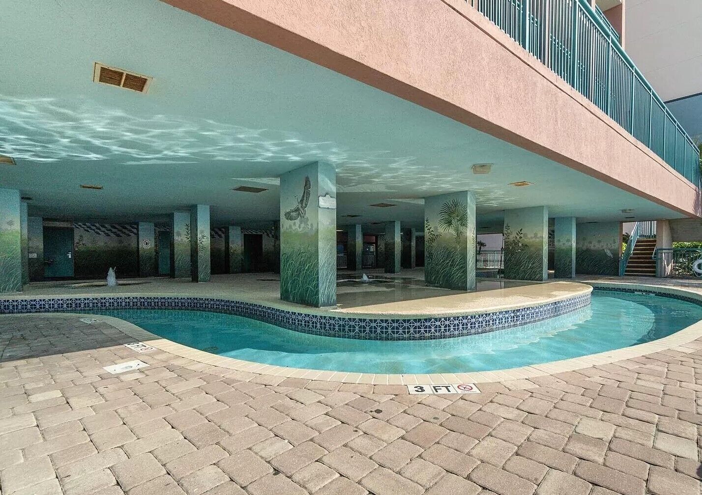 Zona de piscina con borde de azulejos, un camino de ladrillo y un edificio con balcón.