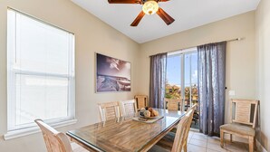A dining area with a glass-top table, wooden chairs, a ceiling fan, and a view through large windows.