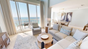 A spacious living room with a large sliding glass door leading to a balcony, a round wooden coffee table, a beige sofa with patterned cushions, and a modern chandelier.