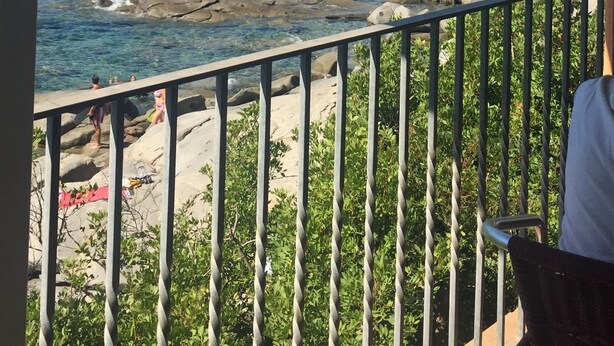 Un balcon offrant une vue sur une zone côtière, avec une eau d’un bleu limpide, un rivage rocheux et une ville lointaine parsemée de bâtiments.