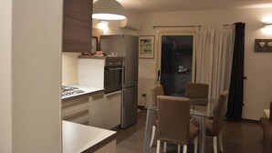 Une cuisine compacte équipée d’une cuisinière à gaz, d’un réfrigérateur et d’un four. On y trouve une table à manger avec des chaises et une cloison en verre. Une grande fenêtre avec des rideaux est visible, et un tableau encadré est accroché au mur.