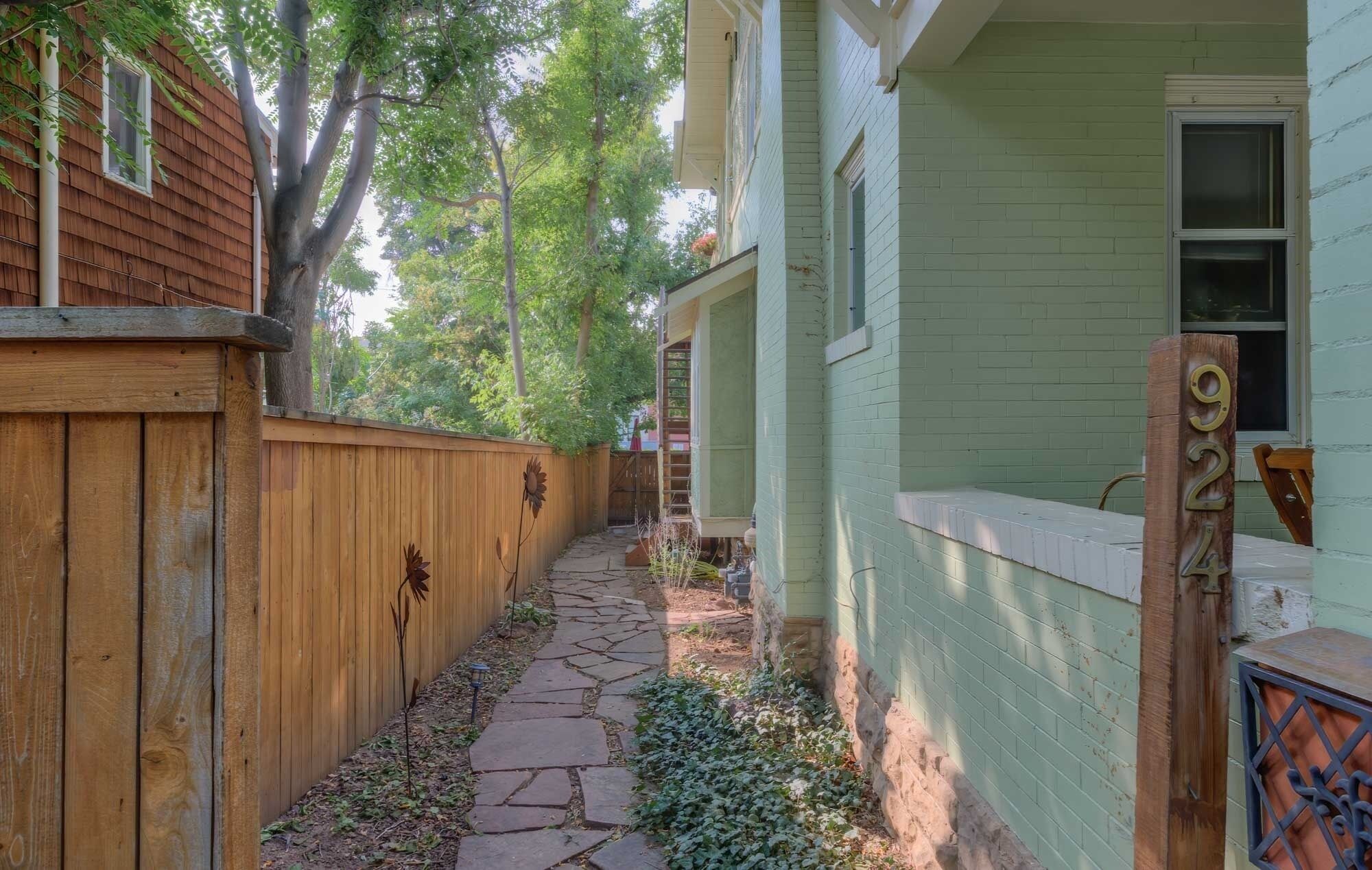 A narrow stone pathway leading to a house with a wooden fence on one side and a green-painted exterior on the other.