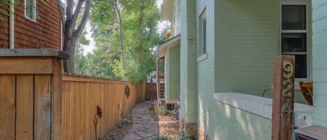 A narrow stone pathway leading to a house with a wooden fence on one side and a green-painted exterior on the other.