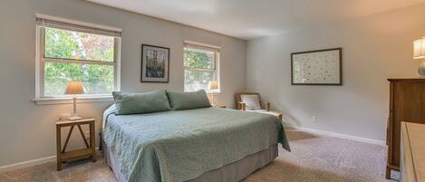 A bedroom with a large bed, two windows, a nightstand, and a lamp.