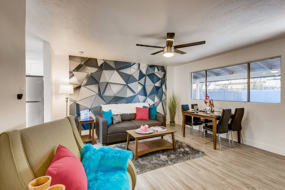 A modern living room with a geometric-patterned wall, a ceiling fan, a dining area with a table and chairs, and a view through large windows.