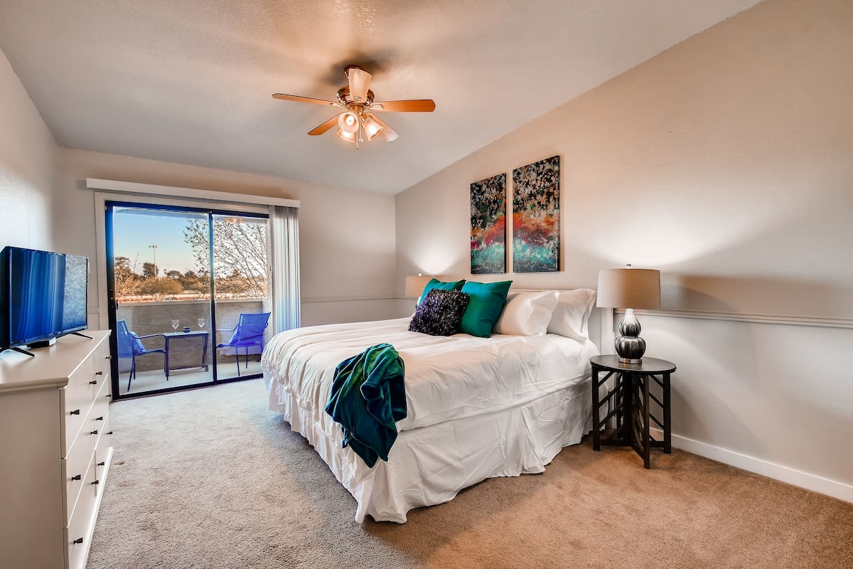 A bedroom with a bed, a ceiling fan, a TV, a lamp, and a view of the outdoors.