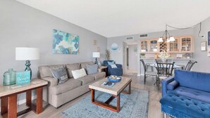 Ein Wohnzimmer mit einer Couch, einem Couchtisch und einem blauen Sessel. Es gibt eine Küchenzeile mit Schränken und einem Esstisch.