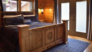A wooden cabin bedroom with a large bed, a nightstand, a lamp, and a view of the outside through a window. - Rowdy Rooster, A Relaxing Cabin 3.4 miles from nearest Broken Bow Lake boat ramp (Broken Bow)