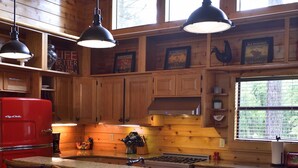 A wooden cabin-style kitchen with a red refrigerator, bar stools, pendant lights, and large windows. - Rowdy Rooster, A Relaxing Cabin 3.4 miles from nearest Broken Bow Lake boat ramp (Broken Bow)