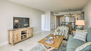 A living room with a television, wooden entertainment unit, and a coffee table with magazines. There is a sofa with patterned cushions and a lamp.