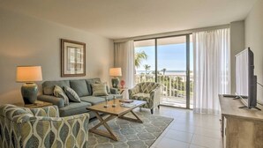 A living room with a sofa, armchairs, a coffee table, and a TV. There is a large window with a view of the ocean.