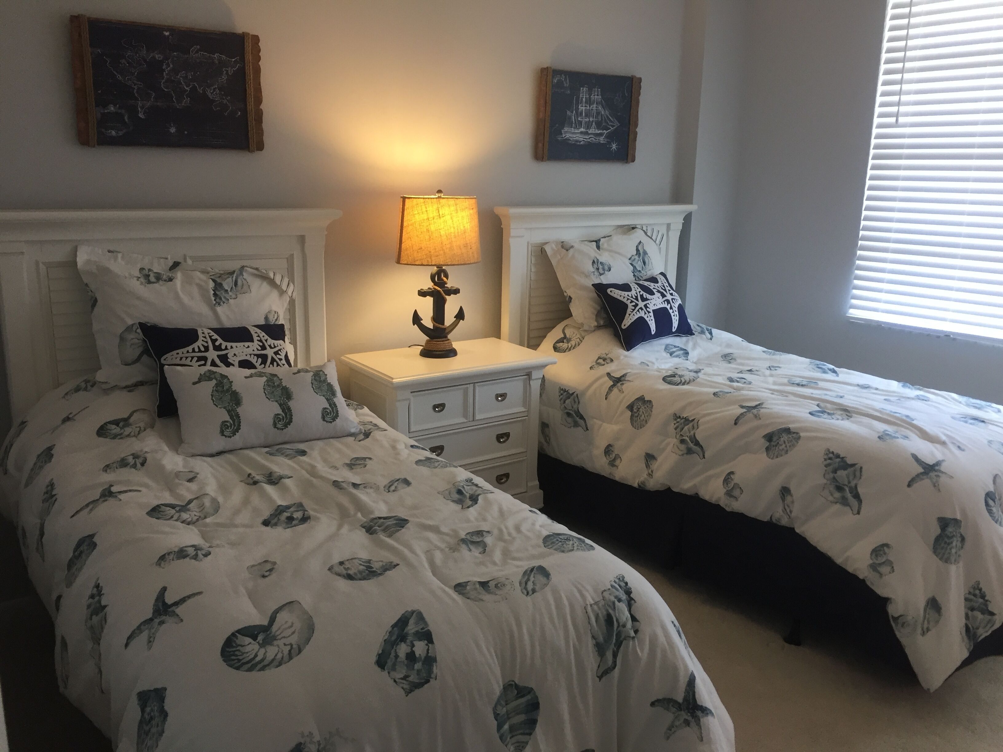 Camera da letto con due letti, ognuno con copriletto a tema nautico, un comodino con lampada e decorazioni murali raffiguranti una mappa del mondo e una nave.