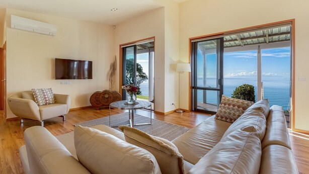 A spacious living room with a large sofa, a coffee table, and a TV mounted on the wall. There is a sliding glass door leading to a balcony with a view of the sea. - Moeraki (Waiheke Island)