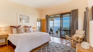A bedroom with a large bed, a chair, a view of the sea, and a balcony. - Moeraki (Waiheke Island)