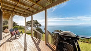 A wooden deck with outdoor seating and a Weber grill, overlooking a coastal view. - Moeraki (Waiheke Island)