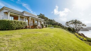 A two-story house with a covered porch, a grassy lawn, and a view of the sea. - Moeraki (Waiheke Island)