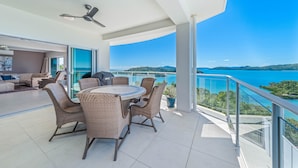 Un balcón moderno con una mesa de comedor y sillas, un ventilador de techo y una vista panorámica del mar y las colinas.