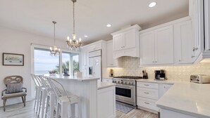 A modern kitchen with white cabinetry, a central island with bar stools, a large stove, and a view of the ocean through a window.