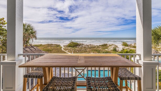 A wooden table with wicker chairs in a covered outdoor area with a view of the ocean and a pool.