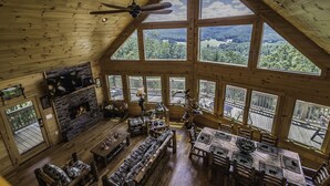 A spacious cabin with a large living area featuring a stone fireplace, wooden furniture, and expansive windows offering a view of the surrounding forest. - A View  To Remember (Morganton)