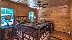 A wooden cabin bedroom with a bed, a ceiling fan, and a window with a view of trees. - A View  To Remember (Morganton)