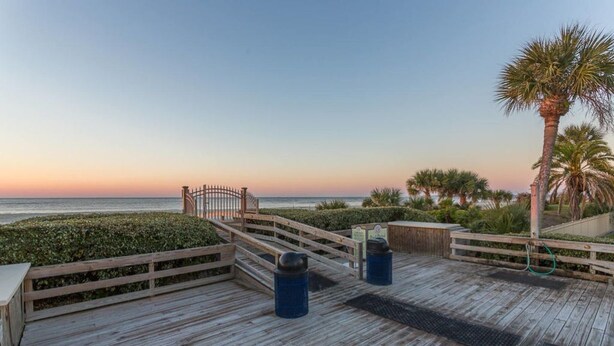A wooden deck with a metal gate, palm trees, and a view of the ocean at sunset. - Top Floor, Partial Oceanview, Peaceful Condo (St. Simons)