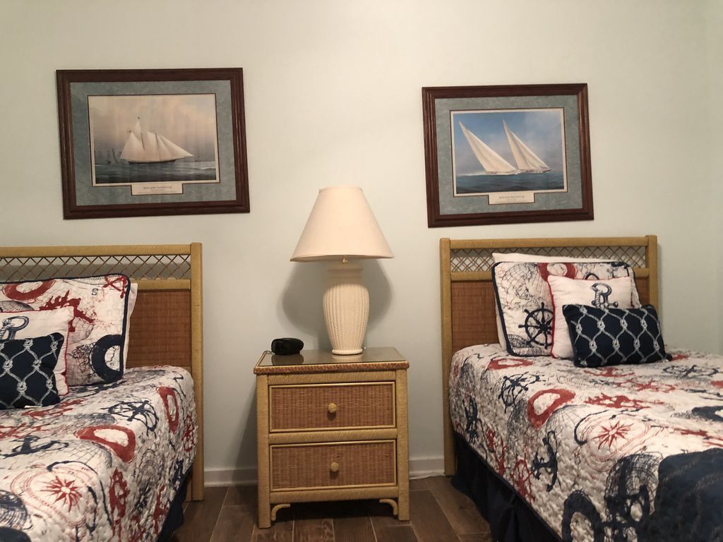 A room with two beds, each with a nautical-themed bedspread, a bedside table with a lamp, and framed pictures of sailboats on the wall.